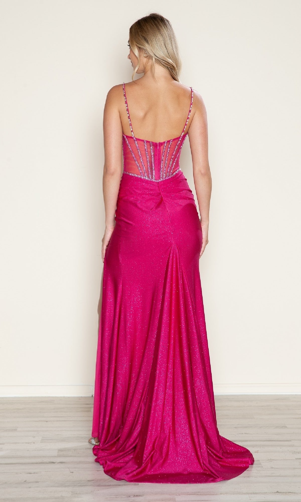A woman stands facing away, wearing the Sheer Beaded-Corset Long Prom Dress (Poly USA 9750), a bright pink, floor-length gown featuring thin straps, glitter detailing, and a partially sheer back.